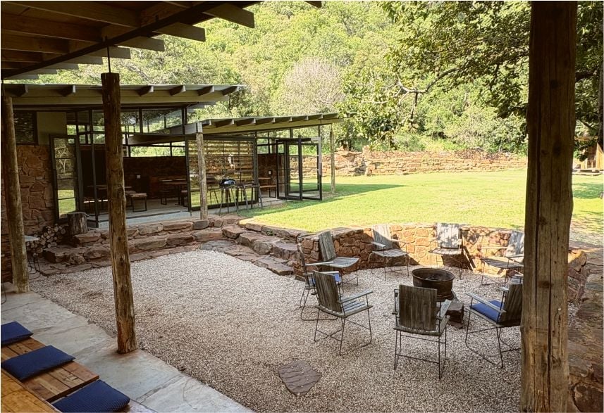 Rustic outdoor patio area with wooden pergola, fire pit, metal chairs, and green lawn with trees in background