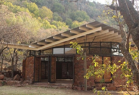 Modern brick structure with wooden pergola roof, dark windows and doors, surrounded by trees and climbing vines