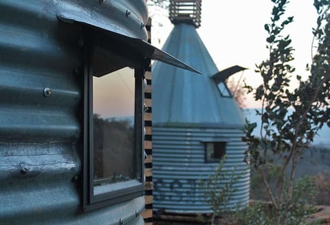 Modern metal cylindrical buildings with corrugated blue-gray siding, featuring large windows and conical roofs in a forested setting