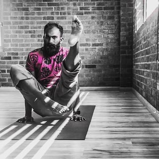 Bearded man in pink shirt sitting on wooden floor against brick wall, leg raised, with selective color photography effect