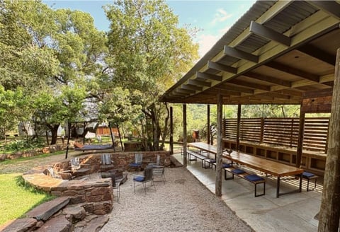 Rustic covered patio with wooden tables overlooking a fire pit and grassy outdoor seating area surrounded by mature trees