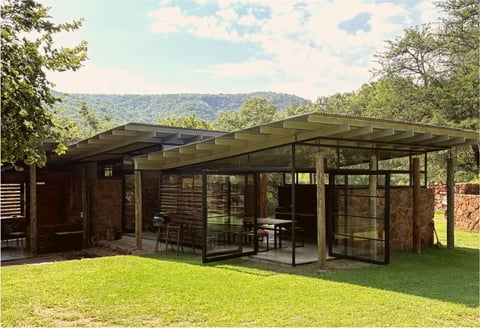 Modern brick and glass pavilion with metal roof on grassy grounds surrounded by forested hills
