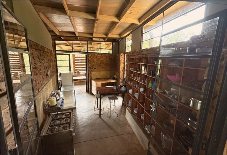 Interior of a rustic workshop or studio with exposed wooden ceiling beams, brick walls, large windows, and dark wooden shelving along the right side
