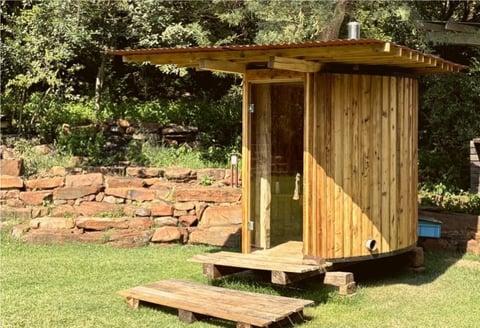 Wooden garden shed with pergola roof and steps on grass lawn, surrounded by trees and stone edging