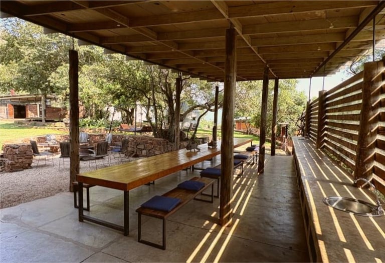 Covered outdoor pavilion with wooden table and benches, surrounded by trees and green grass, with louvered fence on the side