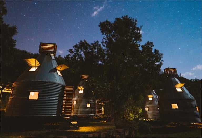 Three illuminated cylindrical structures with glowing windows at dusk surrounded by trees under a starry sky