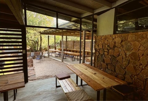 Modern open-air pavilion with wooden tables, benches, natural wood walls, and views of surrounding trees and outdoor seating area