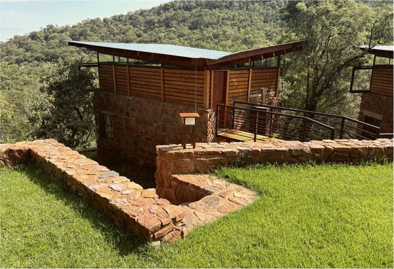 Modern wooden cabin with metal roof overlooking a forested landscape, with a brick fire pit ring in the grassy foreground