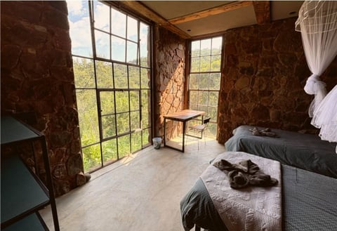 Bedroom interior with large multi-pane windows, dark patterned wallpaper, bed, and vintage furnishings overlooking green landscape