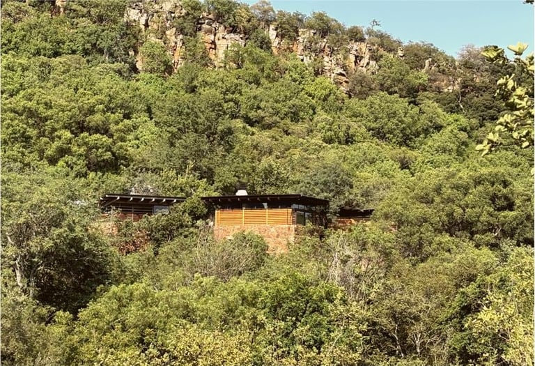 Modern brick building with dark roof nestled among dense green forest and rocky hillside in natural landscape