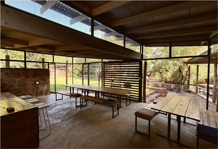 Modern open-concept pavilion with wood beams, large windows, dining tables, and outdoor forest views
