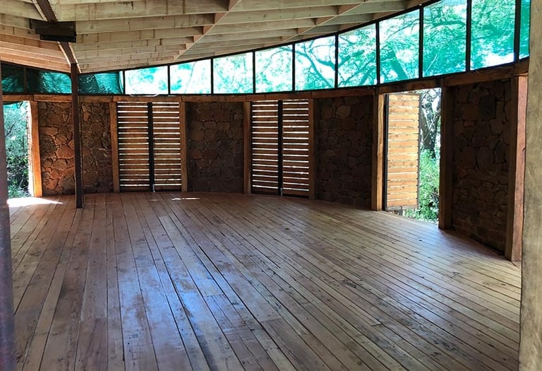 Empty wooden interior space with large windows overlooking greenery, wooden paneling, and louvered doors along the walls