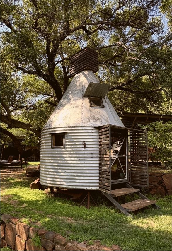Unique corrugated metal cottage with conical roof nestled among large oak trees on a grassy property