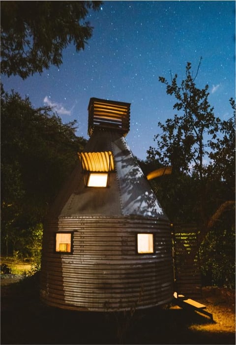 Illuminated cylindrical metal structure with glowing windows at night under starry sky surrounded by trees