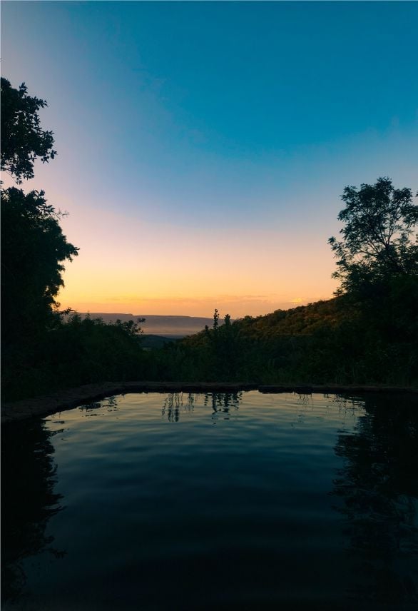 Serene lake at sunset surrounded by silhouetted trees and distant mountains under a gradient sky