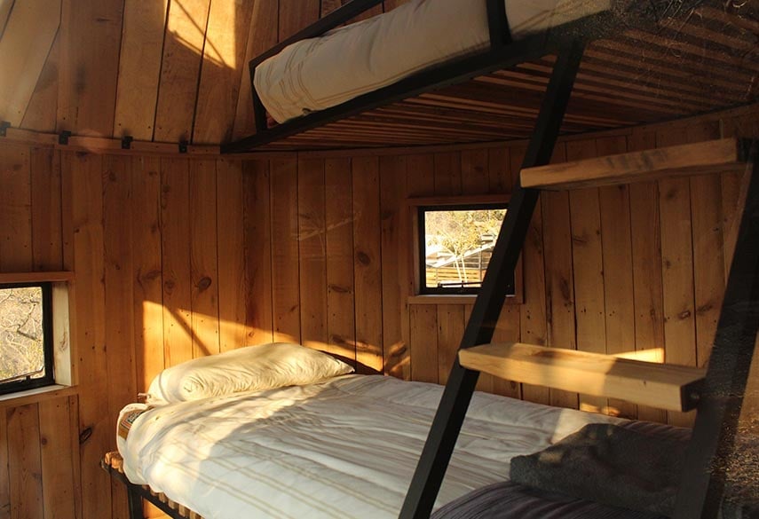 Interior of a wooden cabin bedroom with a bed, small windows letting in sunlight, and exposed wooden beams and walls